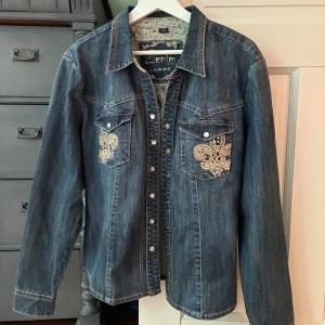 Jeansjacka 🤍⚜️ - säljer en jeansjacka med rhinestones o snygg stitch. jackan har tryckknappar framtill och två fickor. symbol som franksa liljan. slitningarna tillhör design❕ 