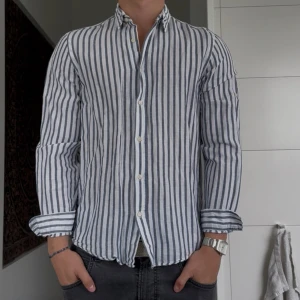 Randig linneskjorta från Zara - Snygg vit och blå randig skjorta från Zara i relaxed fit. Skjortan är tillverkad i linne, har långa ärmar och klassisk krage. Perfekt för dig som gillar en chill och stilren look. Knappar framtill och skön passform.