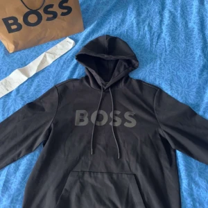 Svart hoodie från BOSS med logga - Svart hoodie från BOSS med stor logga framtill. Klassisk modell med huva, dragsko och känguruficka. Snygg och enkel design, perfekt för dig som gillar stilrena plagg med tydlig branding. Materialet är mjukt och bekvämt, passar till många olika outfits. Storlek L men passar som M.