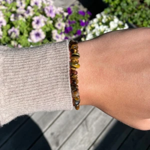 Brunt armband med stenar - Snyggt armband med stenar i bruna och gyllene toner. Stenarna är trädda på en justerbar beige tråd med knyt, vilket gör det enkelt att anpassa storleken. Perfekt accessoar för en naturlig och avslappnad stil.