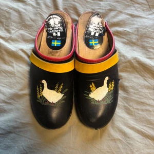 Svenska trätofflor med gåsmotiv - Unika svarta trätofflor med handmålat gåsmotiv och gula detaljer. Sidorna är röda och insidan har svensk flagga. Tillverkade i trä och skinn, med platt sula och klassisk rund tå. Perfekta för dig som gillar traditionell svensk stil med färgglada detaljer. Små skråmor på sidan men i väldigt bra skick