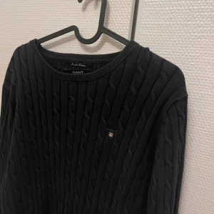 Svart kabelstickad tröja från GANT - Kabelstickad svart tröja från GANT med rund halsringning och diskret broderad logga på bröstet. Tröjan har lång ärm och klassisk passform, perfekt för en stilren och enkel look.