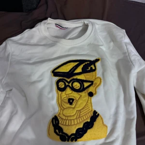 Vit hoodie från Moncler med brodyr - Säljer en clean vit sweatshirt från Moncler i 100% bomull. Tröjan har en stor broderad applikation i gult och svart framtill som verkligen sticker ut. Klassisk rund halsringning och långärmad modell. Perfekt för dig som gillar streetwear med lyxig känsla.