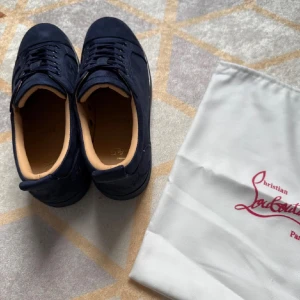 Mörkblå sneakers från Christian Louboutin - Snygga mörkblå sneakers från Christian Louboutin i mocka med klassisk röd sula och diskret logga på sidan. Skorna har rund tå, platt sula och snörning. Insidan är ljusbeige och de kommer med originalpåse. Perfekta för dig som gillar exklusiva detaljer.