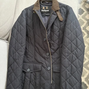 Mörkblå quiltad Barbour jacka - Säljer en mörkblå quiltad jacka från Barbour med klassisk brun krage och snygga knappar. Jackan har dragkedja, två stora fickor framtill och en stilren passform. Perfekt för dig som vill ha en tidlös och stilren look.