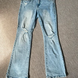 True Religion bootcut jeans ljusblå - Snygga ljusblå bootcut jeans från True Religion, modell Becca Mid Rise. Jeansen har detaljer och hål på knäna, klassiska bakfickor med röd söm och logopatch. Perfekta för en avslappnad och trendig look. Skriv till mig vid minsta fråga. Midjemått: ungefär 42cm rakt över, Innerbenslängden: ungefär 75cm!