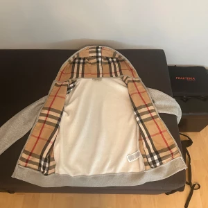 Grå zip-hoodie från Burberry M - Grå hoodie med dragkedja från Burberry, ikoniskt rutigt foder i huvan och insidan. Klassisk broderad logga på bröstet och ribbade muddar. Perfekt för dig som vill ha en stilren och lyxig streetlook med premiumkänsla.