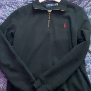 Svart half zip tröja Polo Ralph Lauren - Svart långärmad tröja från Polo Ralph Lauren med klassisk röd broderad logga på bröstet. Tröjan har en snygg half zip-dragkedja vid halsen och är gjord i mjuk bomull. Perfekt för en clean och stilren look. Priset kan diskuteras!