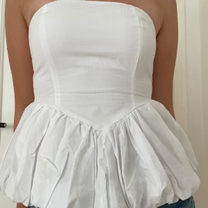 Vit volang tubtopp från Stradivarius - Supersöt vit tubtopp från Stradivarius med volang nertill och smockad rygg för extra komfort. Toppen är axelbandslös och har en snygg, puffig form som ger en trendig look. Perfekt för att skapa en cool och somrig outfit.