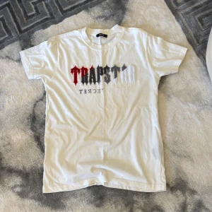 Vit Trapstar t-shirt med tryck - Vit t-shirt från Trapstar med ikoniskt tryck i rött, svart och grått på bröstet. Klassisk rund hals och korta ärmar. Tillverkad i 100% bomull för en skön och avslappnad känsla. Perfekt för dig som gillar streetwear och vill sticka ut.
