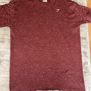 Vinröd Gymshark t-shirt XS - Snygg vinröd t-shirt från Gymshark i storlek XS. T-shirten har ett diskret mönster i tyget och en liten vit logga på bröstet. Tillverkad i ett mjukt och stretchigt material som passar perfekt till träning eller chill. Klassisk rund hals och korta ärmar.