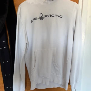 Vit hoodie från Sail Racing - Vit hoodie från Sail Racing med svart logga tryckt över bröstet.                                                                       Pris kan diskuteras 