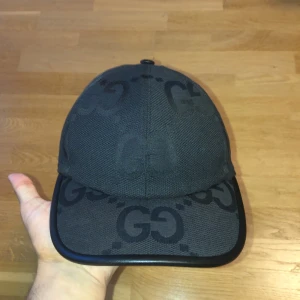 Svart keps från Gucci med GG-mönster - Snygg svart keps från Gucci med klassiskt GG-mönster över hela kepsen. Kepsen har böjd skärm med svart kant i skinn och justerbar rem baktill. Tillverkad i tåligt tyg som ger en exklusiv känsla och passar perfekt till streetwear eller en clean look.