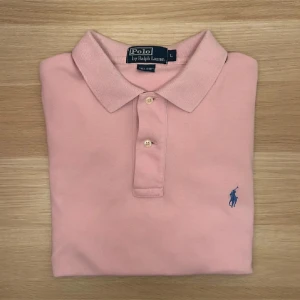 Ralph Lauren pike - Klassisk rosa pikétröja från Ralph Lauren med blå broderad logga på bröstet. Tröjan har korta ärmar, krage och knäppning med två knappar. Tillverkad i mjuk bomull som känns skön mot huden. Perfekt för en clean och stilren look.