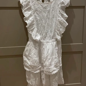 Vit kort klänning med volang och spets - Superfin vit kort klänning med broderade detaljer och volang längs axlarna. Köpt till studenten men användes aldrig!