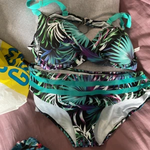 Tropisk bikini med palmmönster - Färgglad bikini med tropiskt palmmönster i svart, turkos, lila och vitt. Överdelen har vadderade kupor och justerbara axelband i turkos. Underdelen har dekorativa band framtill för extra detalj.