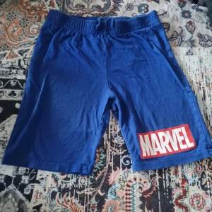 Blå Marvel shorts från H&M - Snygga blå shorts från H&M med Marvel-logga i vitt och rött på ena benet. De har elastisk midja för skön passform och är gjorda i mjuk bomull. Perfekta för dig som gillar superhjältar och vill ha en avslappnad stil.