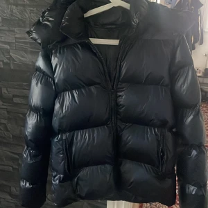 Svart pufferjacka från Everest - Säljer en svart pufferjacka från Everest med glansig finish och stor huva. Jackan har dragkedja framtill och två fickor med dragkedja. Perfekt för kalla dagar när du vill hålla dig varm och ändå se snygg ut.