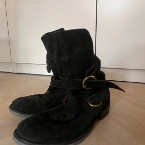 Svarta mockaboots med spännen - Snygga svarta boots i mocka med coola mässingsfärgade spännen och brunt skinnfoder. De har en platt sula och rund tå, perfekt för dig som gillar en avslappnad men edgy stil. Passar till både jeans och kjol för en trendig look.