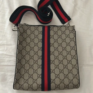  axelväska med mönster - Snygg axelväska  i beige canvas med klassiskt mönster och bred axelrem i rött och marinblått. Väskan har en dragkedjeficka på framsidan och svarta skinn-detaljer. Perfekt för dig som vill ha en ikonisk och trendig accessoar.