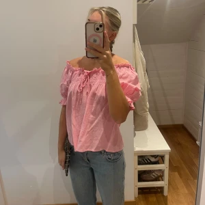 Rosa offshoulder blus med volang - Söt rosa blus med offshoulder och korta puffärmar. Smockad upptill med volangkant och knyt framtill. Luftig och lätt i bomull, perfekt för varma dagar. Passar till både jeans och kjol för en trendig look.