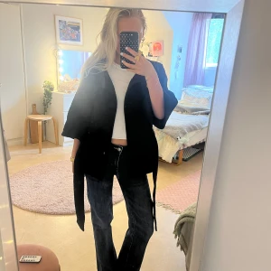 Svart kappa med bälte och stora knappar - Säljer en svart kappa med trekvartsärm och bälte i midjan. Jackan har stora tygklädda knappar framtill och ett strukturerat tyg som ger en cool känsla. Perfekt för dig som gillar stilrena och tidlösa plagg med en twist.