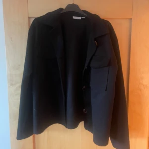 Svart overshirt med fickor och knappar - Storlek M men passar mig som vanligtvis har S på jackor. Stilren svart overshirt med klassisk krage, stora bröstfickor och knäppning framtill. Jackan har en rak passform och är tillverkad i ett mjukt tyg som känns skönt mot huden. Perfekt att slänga över en hoodie eller t-shirt för en chill look.
