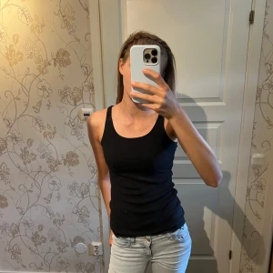 Svart linnetopp med spetsdetaljer - Svart linnetopp med smala axelband och snygga spetsdetaljer både i ryggen och längst ner vid fållen. Toppen har en tight passform och knappar i ryggen som extra detalj. Perfekt att styla med jeans eller kjol för en cool look.