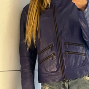 Lila/mötkblå skinnjacka från Estelle, stl L - Snygg lila skinnjacka från Estelle med coola detaljer som fyra dragkedjefickor framtill och spänne i midjan. Jackan har en rak passform, lång ärm och stilren krage. Perfekt för dig som vill sticka ut med en färgstark look.