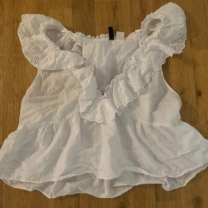 Vit volangblus från H&M Divided - Supersöt vit blus från H&M Divided med breda volanger och broderade spetsdetaljer längs axlarna. Blusen är ärmlös och har en luftig, flowy passform med volangkant nertill. Perfekt för en somrig look och lätt att matcha med jeans eller kjol.