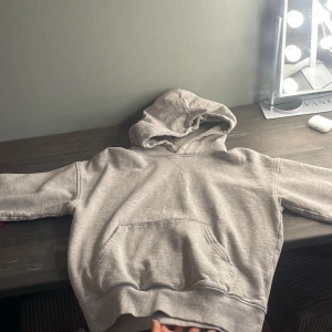 Grå hoodie med huva och magficka - Säljer en enkel och stilren grå hoodie med huva och stor magficka. Tröjan har långa ärmar och ribbade muddar vid ärmslut och nederkant. Perfekt för chill dagar eller när du vill ha något bekvämt och snyggt.