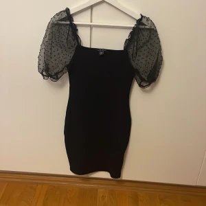 Svart kort klänning med puffärmar Amisu - Supersnygg svart kort klänning från Amisu med transparenta puffärmar i mesh och små prickar. Tight passform och fyrkantig ringning ger en trendig look. Perfekt för dig som vill sticka ut med coola detaljer och volym på ärmarna.