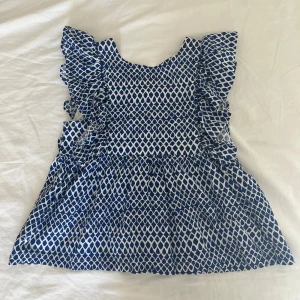 Blåvit mönstrad blus med volangärmar - Supersöt blus från Mango i fin kvalitet med 89% bomull och 11% linne. Blusen är blåvit med snyggt mönster och volangärmar. Smockad rygg för extra komfort och stretch. Perfekt för varma sommar dagar. Blusen är i storlek 152 och passar på mig som är 173 cm lång och har en storlek xs, men skulle däremot säga att den passar bättre på någon med storlek xxs. Blusen har aldrig kommit till användning och är därför i nyskick.