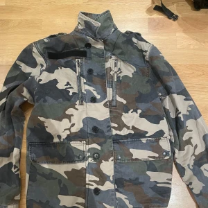 Camouflagejacka från Zadig & Voltaire - Snygg camouflagejacka från Zadig & Voltaire med coola detaljer som stora fickor, knappar och hög krage. Jackan har ett militärinspirerat mönster i grönt, beige och brunt. Perfekt för dig som vill sticka ut med en streetig look.