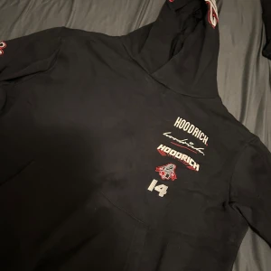 Svart hoodie från Hoodrich med tryck - Svart hoodie från Hoodrich med stora broderade och tryckta detaljer i vitt och rött på bröst, rygg, ärm och huva. Hoodien har huva, känguruficka och coola sportiga loggor som sticker ut. Perfekt för dig som gillar streetwear vibes.