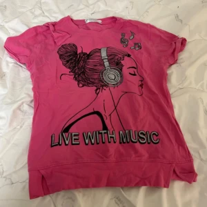 Rosa t-shirt med musiktryck - Cool rosa t-shirt med tryck av en tjej med hörlurar och texten 'LIVE WITH MUSIC' framtill. T-shirten har korta ärmar och rund hals. Perfekt för dig som älskar musik och vill sticka ut med en färgstark stil. Använd bara en gång men super fin. 