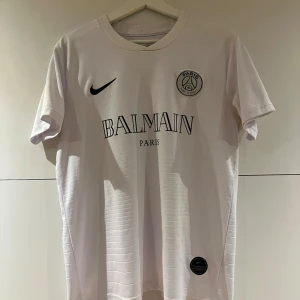 Vit PSG x Balmain fotbollströja Nike - Snygg vit fotbollströja från Nike i samarbete med PSG och Balmain. Tröjan har korta ärmar, rund halsringning och tryckta loggor på bröstet. Materialet är lätt och andas, perfekt för match eller träning. Stilsäker design med exklusiv känsla.