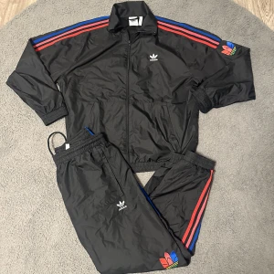 Svart Adidas tracksuit med färgglada detaljer - Säljer en svart Adidas tracksuit med klassiska röda och blå ränder längs ärmar och ben. Setet har broderad Adidas-logga på bröst och ben samt färgglad trefoil-detalj på ärm och byxben. Jackan har dragkedja och resår i ärmslut och midja.