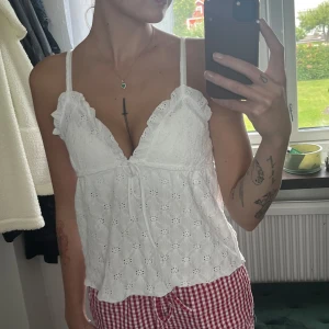 Vit broderad linnetopp med volang - Supersöt vit linnetopp från 157 med broderade blommor, smala axelband och volangdetaljer vid ringningen. Toppen har v-ringning och knyt framtill, perfekt för sommarens varma dagar. Lätt och luftig känsla.