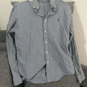 Rutig skjorta från Polo Ralph Lauren - Snygg blåvit rutig skjorta från Polo Ralph Lauren i slim fit-modell. Skjortan har klassisk krage, långa ärmar och knappar framtill. Tillverkad i bomull och prydd med den ikoniska röda loggan broderad på bröstet. Perfekt för en stilren och avslappnad look.