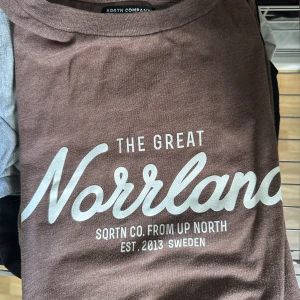 Brun t-shirt The Great Norrland - Brun t-shirt från SQRTN Company med trycket 'The Great Norrland' i vitt framtill. Använd 2 gånger 