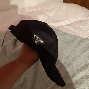 Svart keps från Prada i bomull - Snygg svart keps från Prada med klassisk böjd skärm och justerbart spänne bak. Kepsen har Pradas ikoniska metallogo på sidan och är tillverkad i 100% bomull. Perfekt accessoar för en clean och lyxig look.