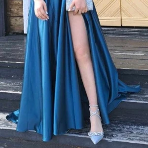 Jägargrön klänning med slits - Använd bara i några timmar, så den är i nyskick. Lyxigt material och sitter jättefint. Klänningen har ett passande spetsutsmyckat livstycke med ömtåliga spaghettiband och en snörning bak för en justerbar passform. Den A-linjede satinkjolen flyter elegant med en golvlång fåll, vilket ger sofistikering. Kombinationen av strukturerade detaljer och en flödande siluett skapar en raffinerad och graciös look.