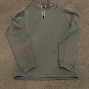 Grå half zip tröja från FootJoy - Snygg grå långärmad tröja från FootJoy med half zip och hög krage. Perfekt för lager-på-lager och har en stilren look med diskret FJ-logga på kragen och ryggen. Tillverkad i mjukt och stretchigt material för extra komfort.
