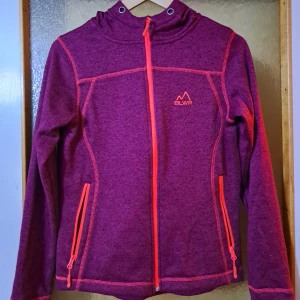 Lila fleecejacka med huva från BLWR - Säljer en lila fleecejacka från BLWR med huva och snygga orange detaljer. Jackan har dragkedja framtill och på fickorna, samt långa ärmar. Stilren och sportig look. Storlek S, men passar för M också.
