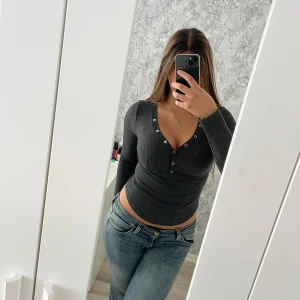 Mörkgrå ribbad långärmad topp - Säljer en mörkgrå ribbad långärmad topp med v-ringning och dekorativa knappar framtill. Modellen är figurnära och croppad, samt lite kortare på sidorna av magen än främre magen och ryggen. Skön stretchig bomullsblandning som sitter snyggt på kroppen.