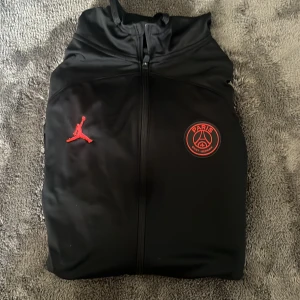 Svart PSG x Jordan tröja, slim fit M - Svart tröja  från Jordan i samarbete med Paris Saint-Germain. Slim fit-modell med hel dragkedja, röda loggor på bröstet och sportig look. Perfekt för dig som gillar streetwear.