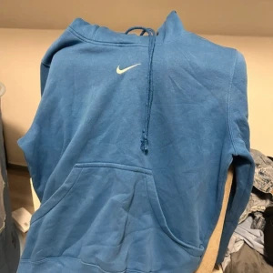 Blå Nike hoodie med huva - Blå hoodie från Nike med klassisk vit swoosh-logga på bröstet. Tröjan har huva med dragsko och en stor känguruficka framtill. Tillverkad i mjuk bomullsmix, perfekt för chill eller streetwear. Har en fläck kan gå bort i tvätten, tvättar såklart innan