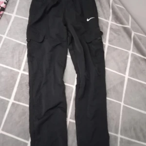 Svarta cargobyxor från Nike - Svarta cargobyxor från Nike med klassisk vit swoosh-logga på benet. Byxorna har elastisk midja, knappstängning och stora fickor på sidorna. Perfekta för en sportig och avslappnad stil. Materialet känns lätt och smidigt.