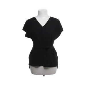 Svart omlottopp med knytband - Stilren svart omlottopp med v-ringning och knytband i midjan. Toppen har korta ärmar och en avslappnad passform som ger en snygg siluett. Perfekt för dig som gillar minimalistisk stil och vill ha något enkelt men ändå trendigt.